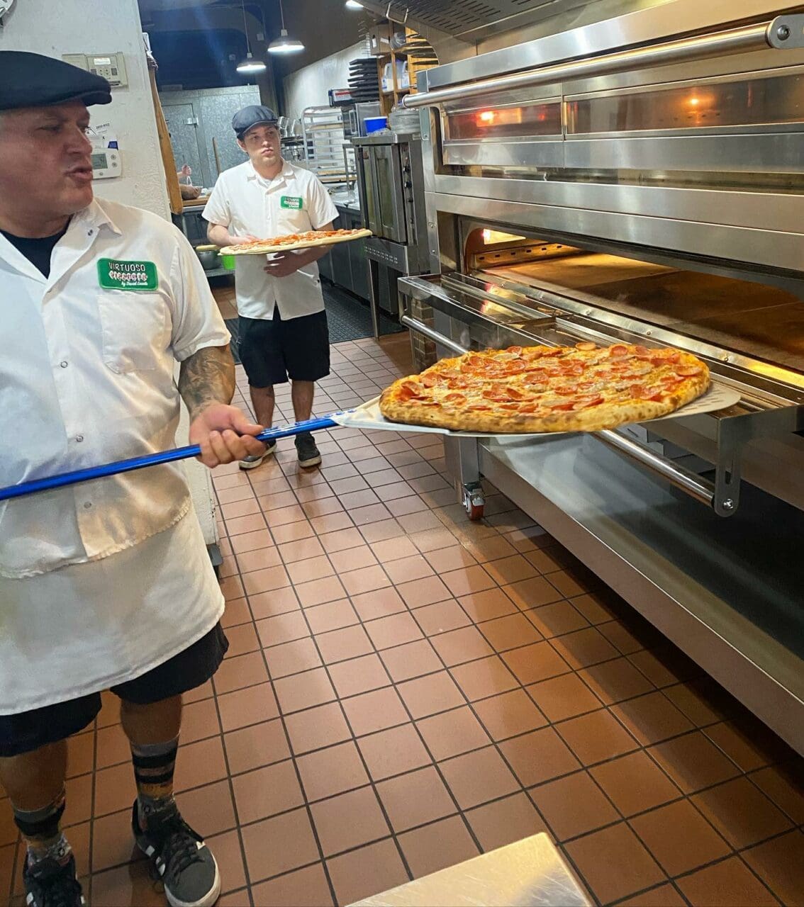 a man with a hat, whit chef's coat, and shorts putting a pizza in the oven.