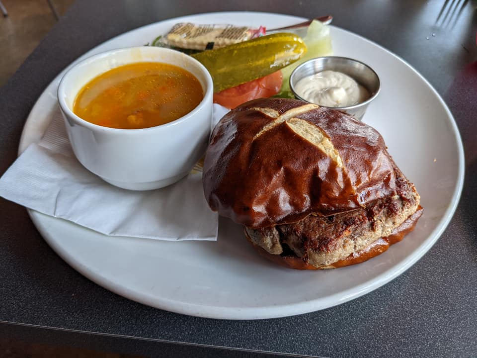 a white plate with a hamburger, soup, and garnishments. 
