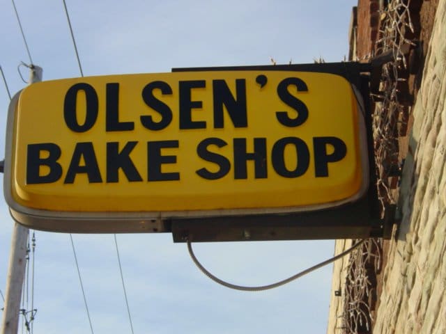 a yellow sign hanging off the side of a building and it says Olsen's Bake Shop