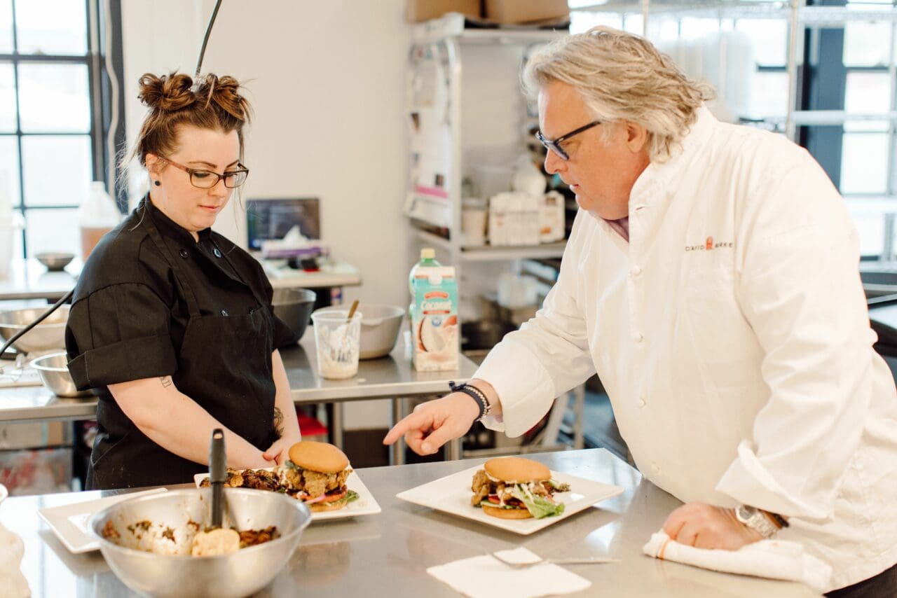 a female chef wearing a black chef's smock and a male chef wearing a white chef's smock looking over several vegan dishes on a steal table.