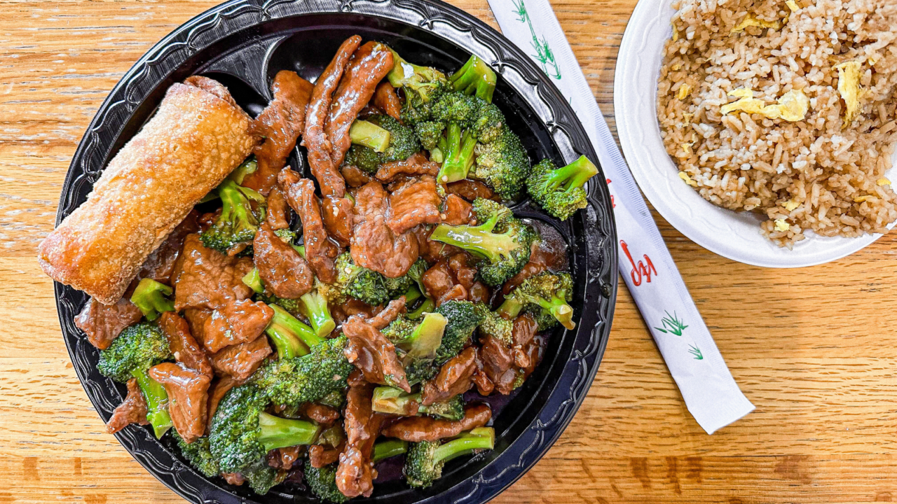 a plate of broccoli beef, fried rice, and an egg roll sitting on a butcher block table