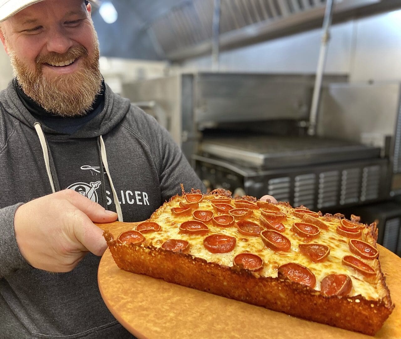 a bearded man in a gray hoodie holding a pepperoni pizza Detroit Deep Dish style.
