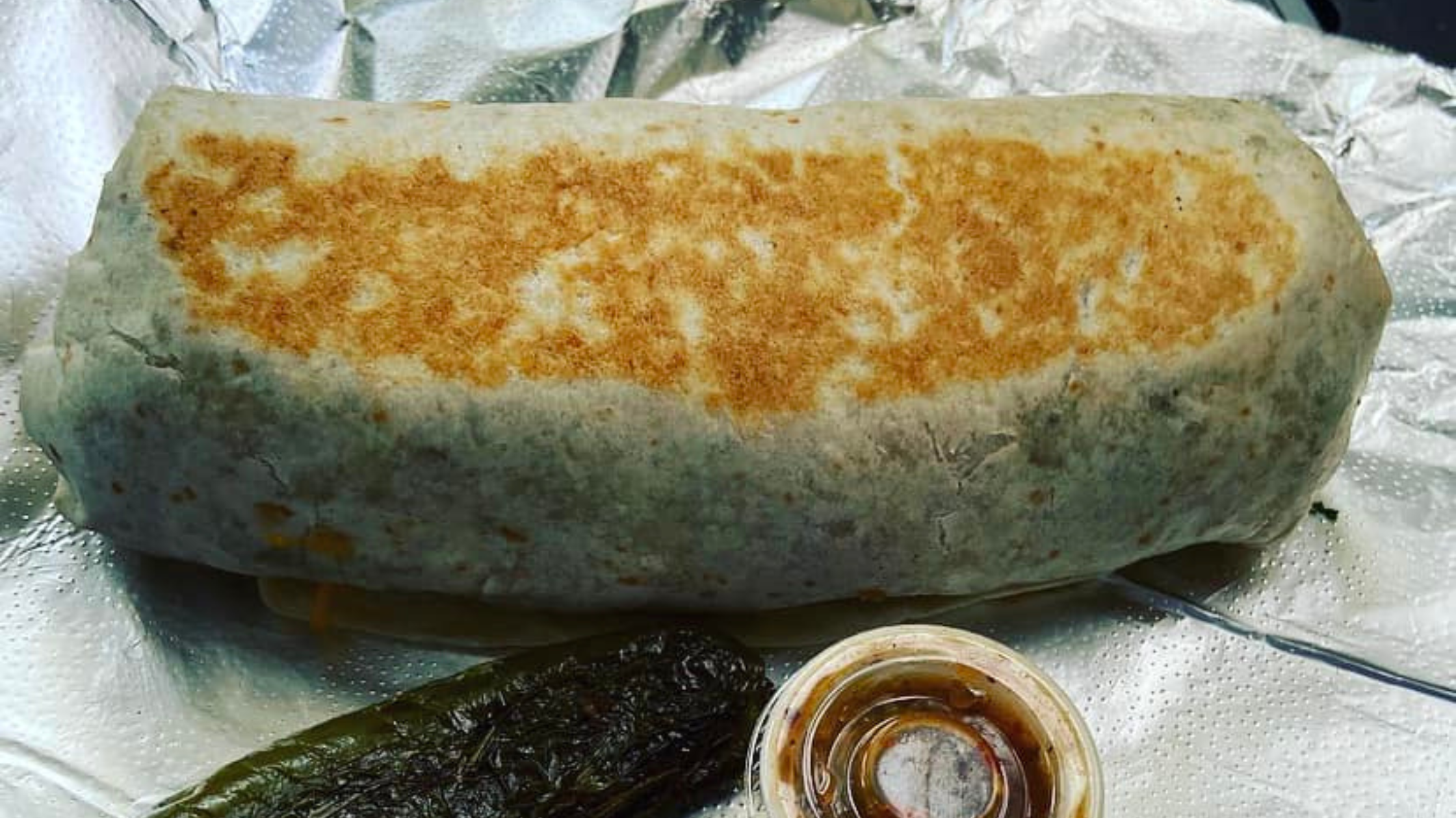 A golden, grilled breakfast burrito from The Churro Spot in Omaha, served with a roasted jalapeño and side of house-made salsa on foil.