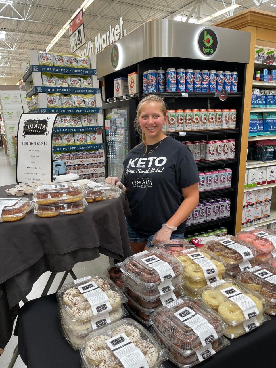 young woman wearing an omaha bakery tee standing next to a display of keto donuts at hy-vee
